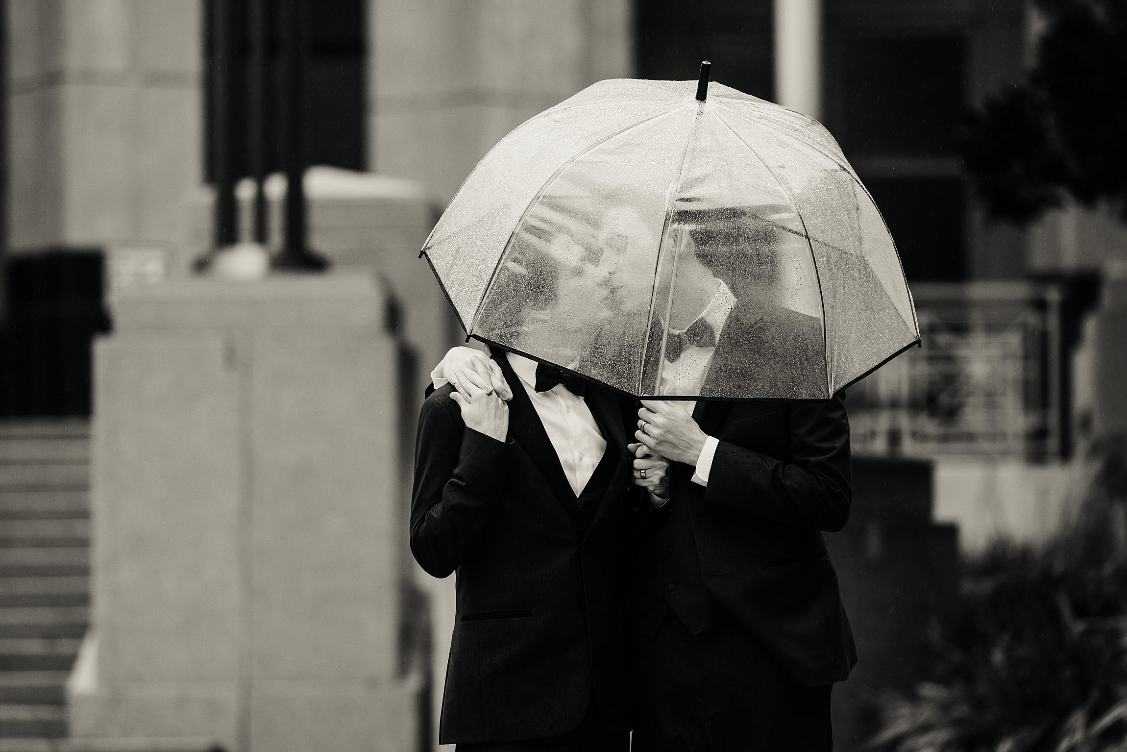 a gay couple hides under a clear umbrella in the rain after their orange county courthouse wedding in orlando