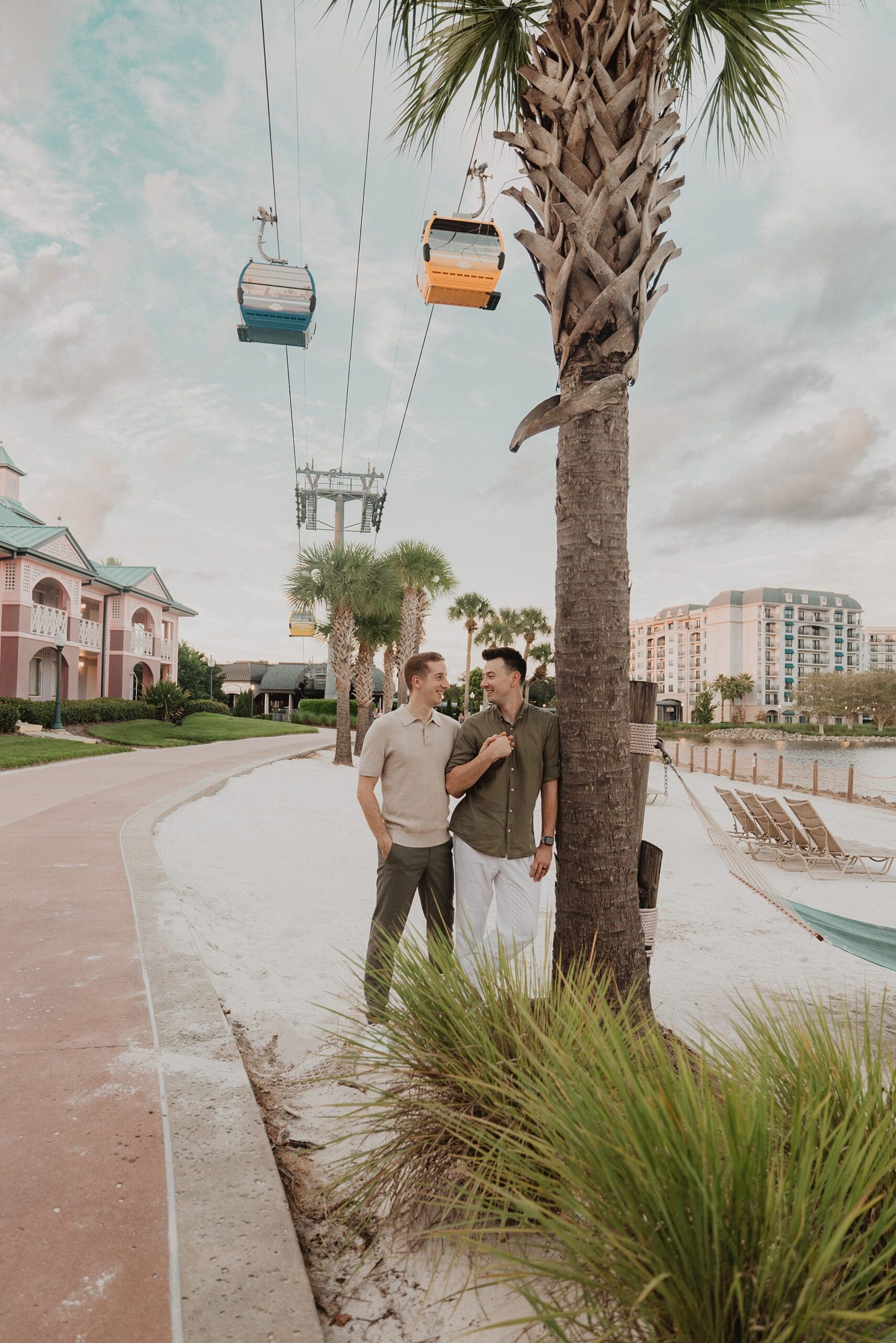 LGBTQIA+ elopement portrait of two grooms wearing casual tan and brown outfits on the beach at an Orlando Florida resort, with cable car trams visible above, captured by Gracie May Photography