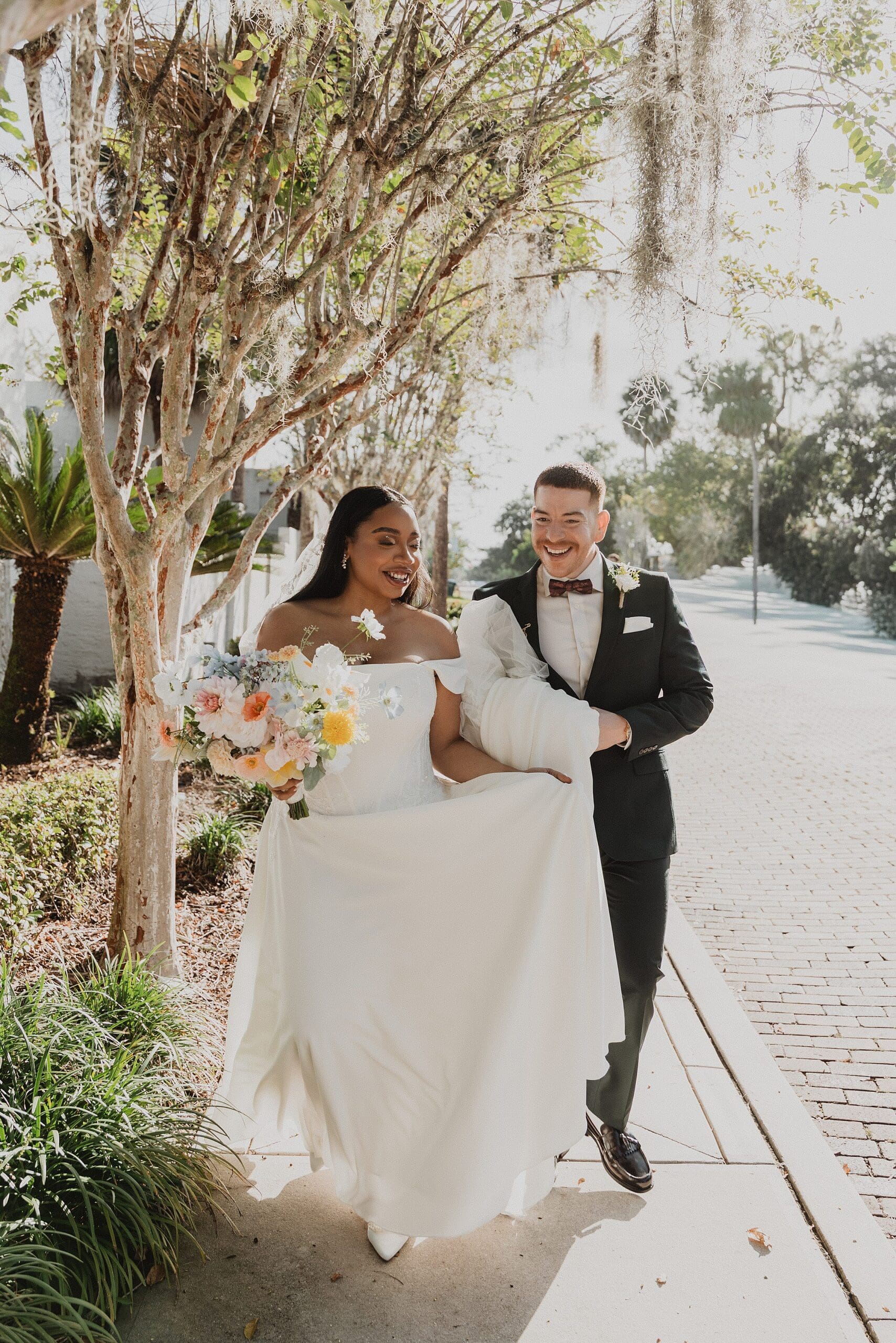 candid couple portrait of a bride holding a bouquet and a groom holding her white gown's train while walking on a brick path framed by trees at a micro wedding at Maitland Art and History Center captured by Gracie May Photography