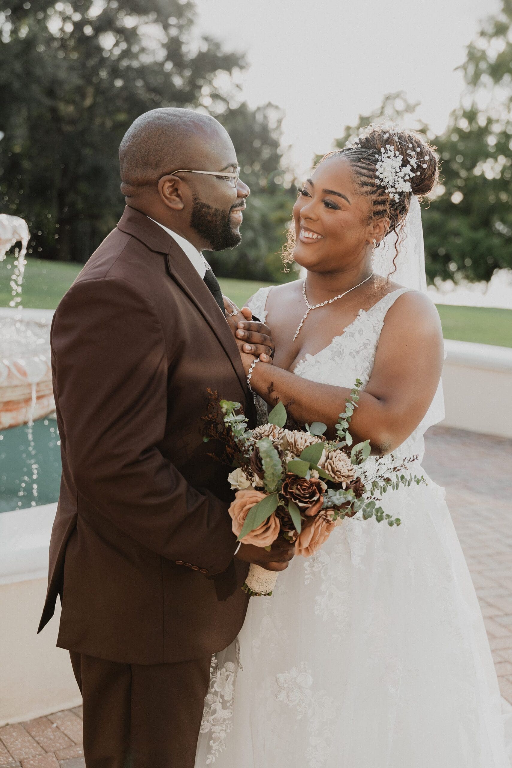 bride wearing white and groom wearing a chocolate brown suit embrace in front of a tiered fountain on the lawn at Lake Mary Events Center in Florida, captured by Gracie May Photography