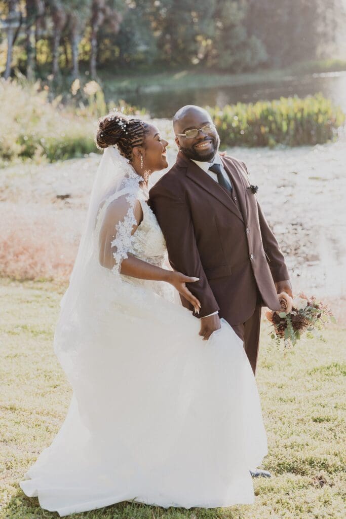 bride wearing white and groom wearing a chocolate brown suit laugh and walk on the waterfront lawn at Lake Mary Events Center in Florida, captured by Gracie May Photography