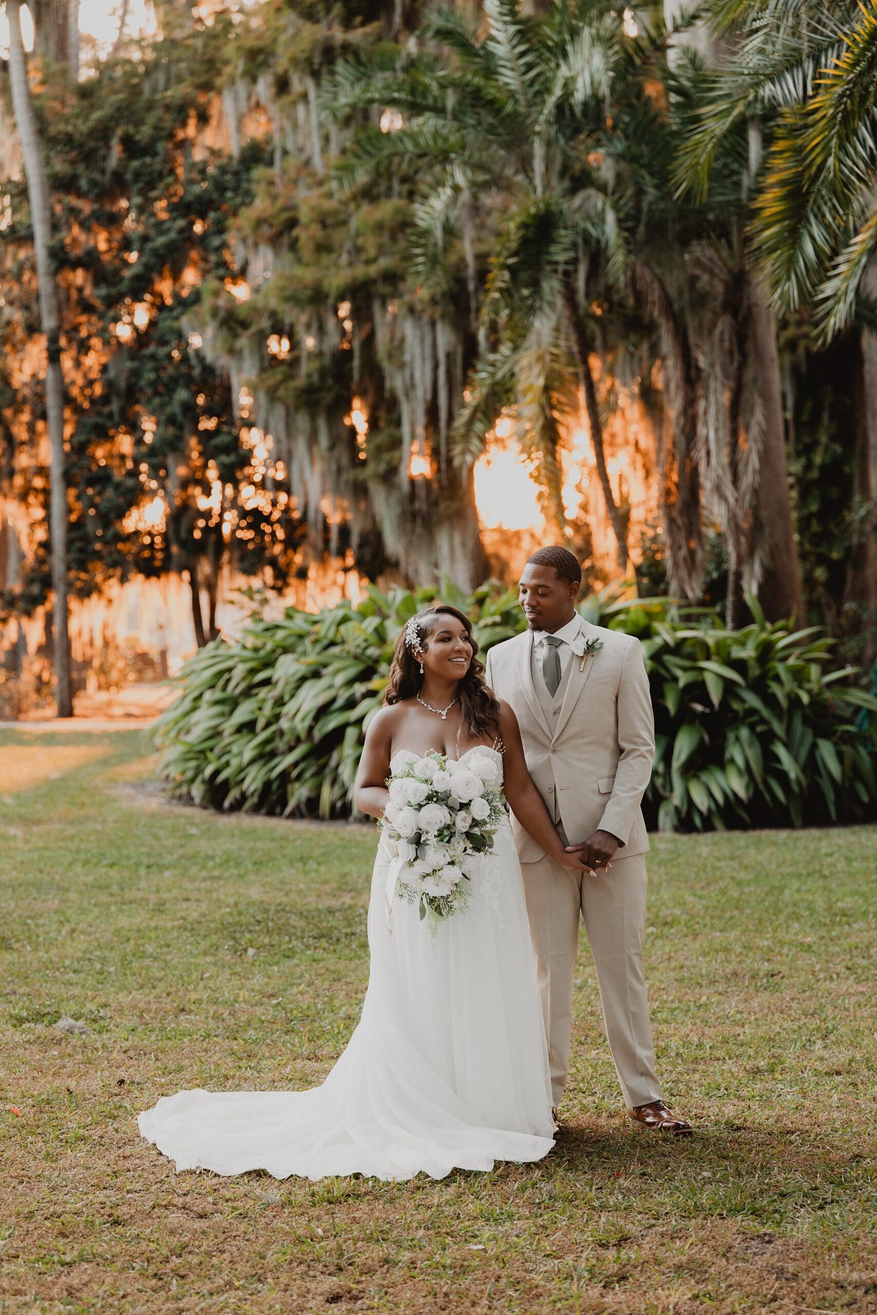 bride and groom hold hands in front of trees with orange sunset golden hour sunlight glowing through at Kraft Azalea Garden in Winter Park, Florida, captured by Gracie May Photography