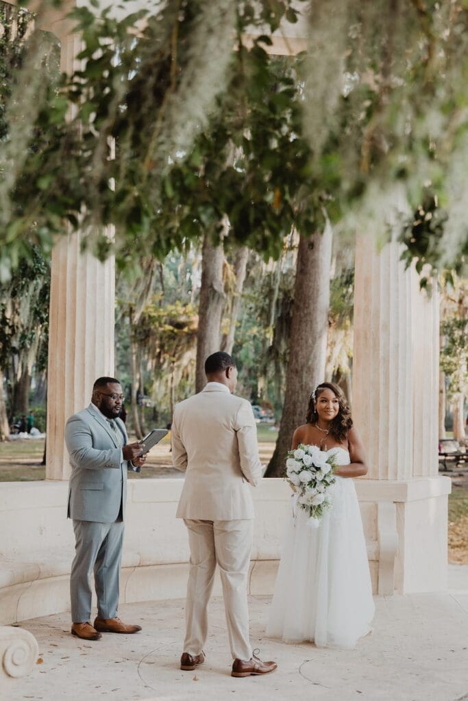 wide shot of bride and groom and officiant at the Exedra Monument during their sunset golden hour micro wedding ceremony at Kraft Azalea Garden in Winter Park, Florida, captured by Gracie May Photography