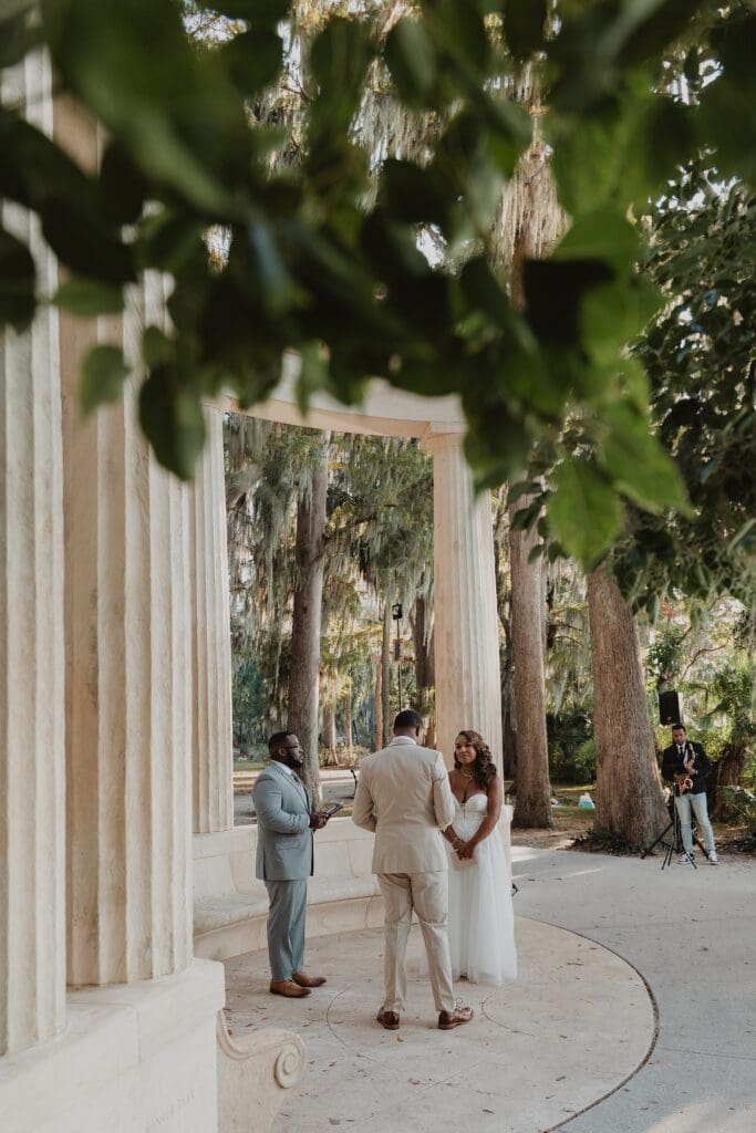 wide shot of bride and groom and officiant at the Exedra Monument during their sunset golden hour micro wedding ceremony at Kraft Azalea Garden in Winter Park, Florida, captured by Gracie May Photography
