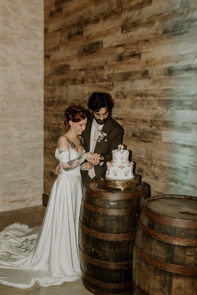 groom in brown suit and bride in an off-the-shoulder gown but their wedding cake on a whiskey barrel at this vintage style rainy day wedding at The Venue at Gentry Pines, captured by Gracie May Photography