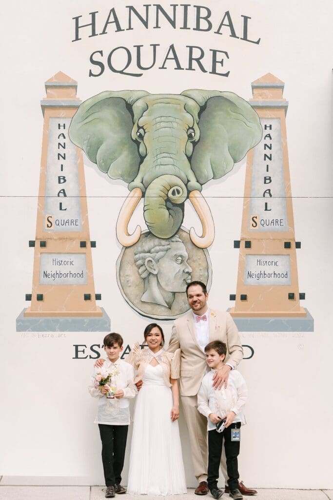 bride in a white dress with a Filipino style jacket and groom in a tan linen suit pose with their sons by a Hannibal Square mural in Winter Park Florida, captured by Gracie May Photography