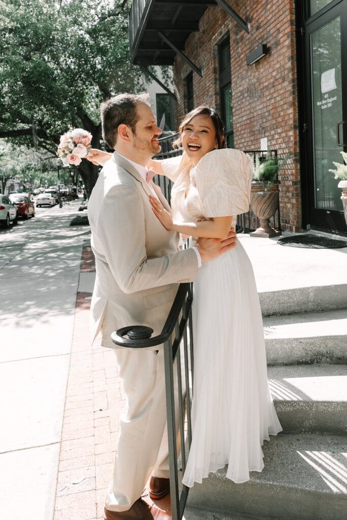 bride in a white dress with a Filipino style jacket and groom in a tan linen suit laugh and embrace on the sidewalk in Winter Park Florida, captured by Gracie May Photography