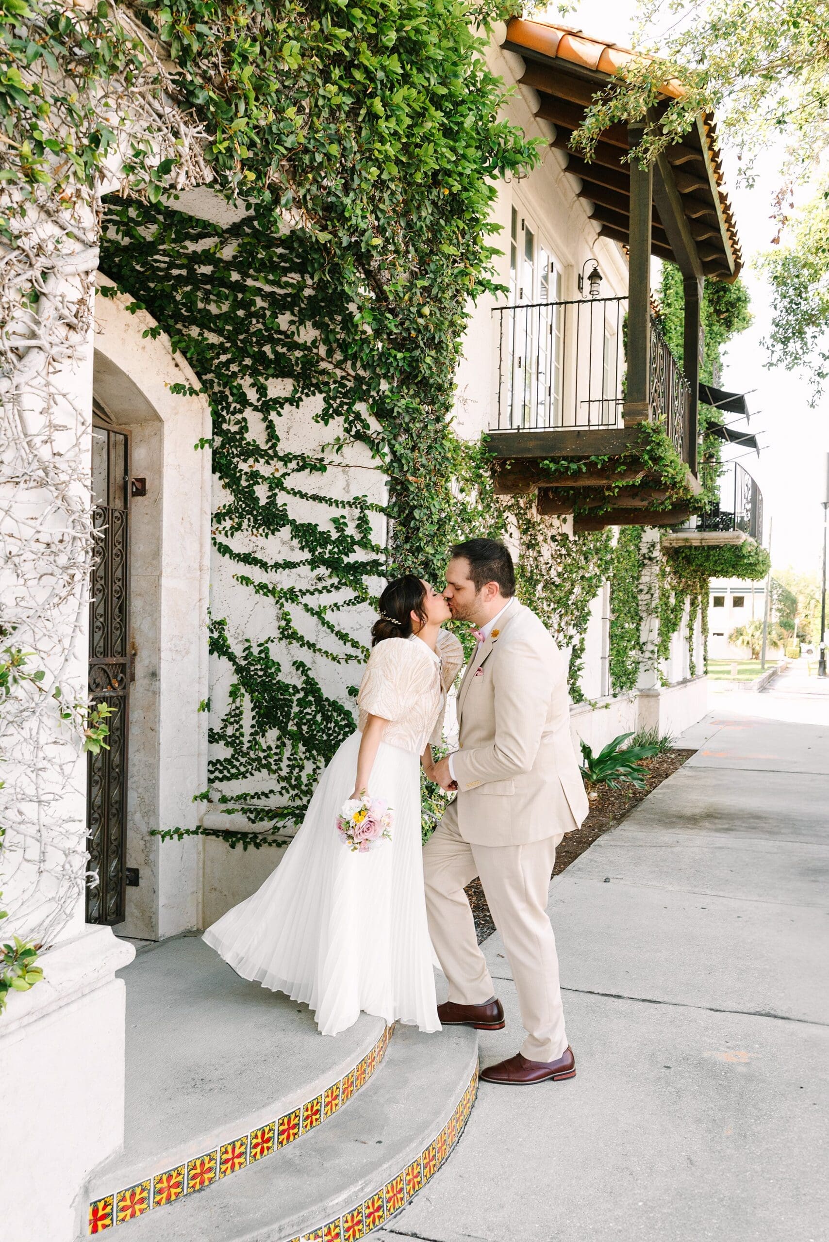 bride in a white dress with a Filipino style jacket and groom in a tan linen suit kiss on the sidewalk in Winter Park Florida, captured by Gracie May Photography