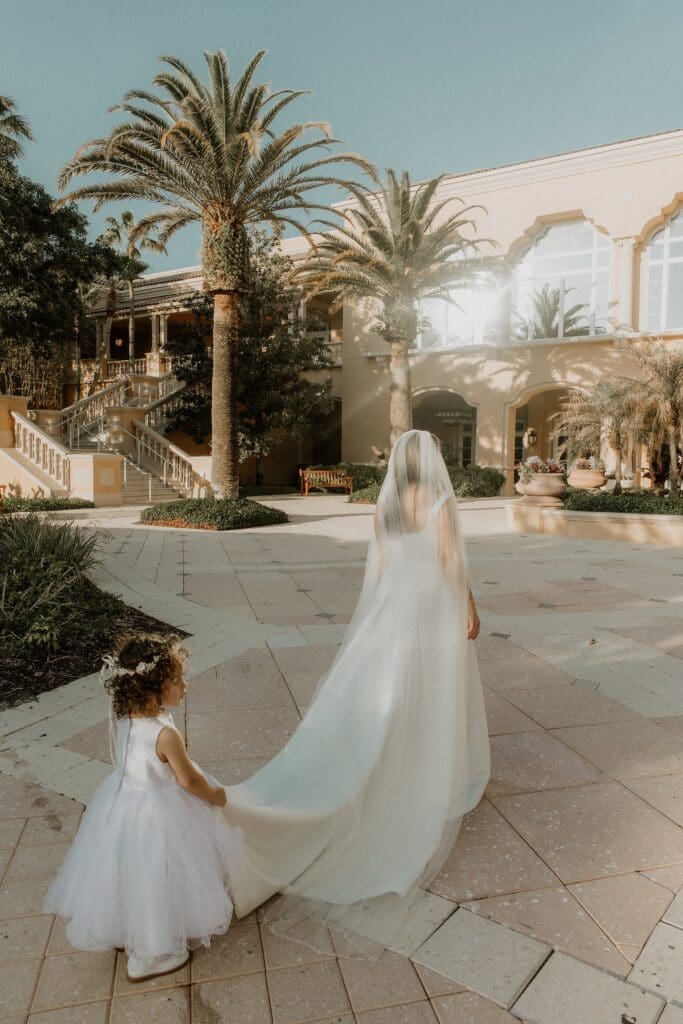 a color photo of a young daughter carrying her mom's wedding dress train as they walk toward daddy for their wedding first look
