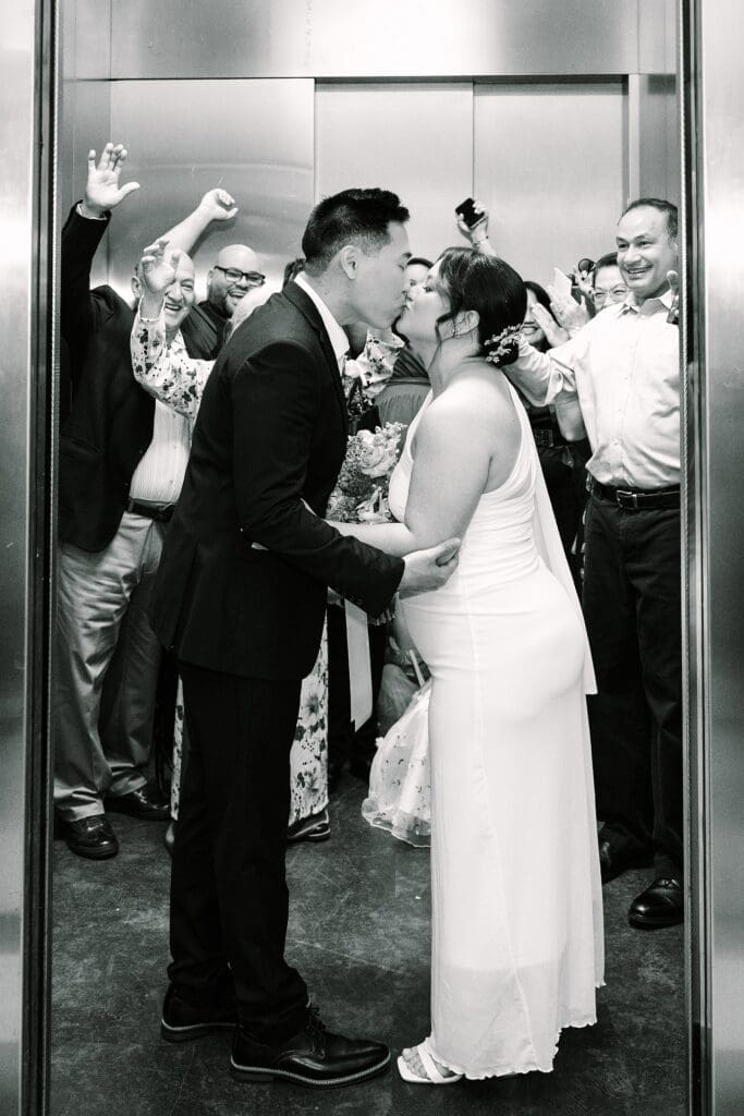 black and white flash shot of a bride and groom in formal wedding attire  kissing in front of their family in the elevator at the Orange County Courthouse in Orlando, captured by Gracie May Photography.