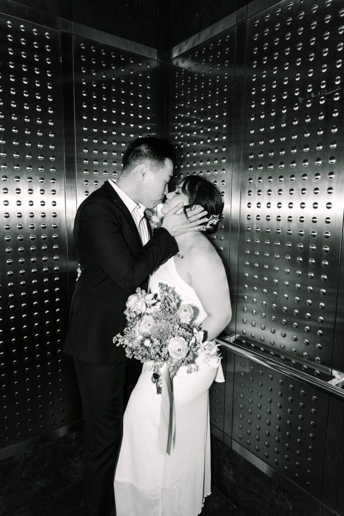 black and white flash shot of a bride and groom in formal wedding attire with a bright spring bouquet kissing in the elevator at the Orange County Courthouse in Orlando, captured by Gracie May Photography.