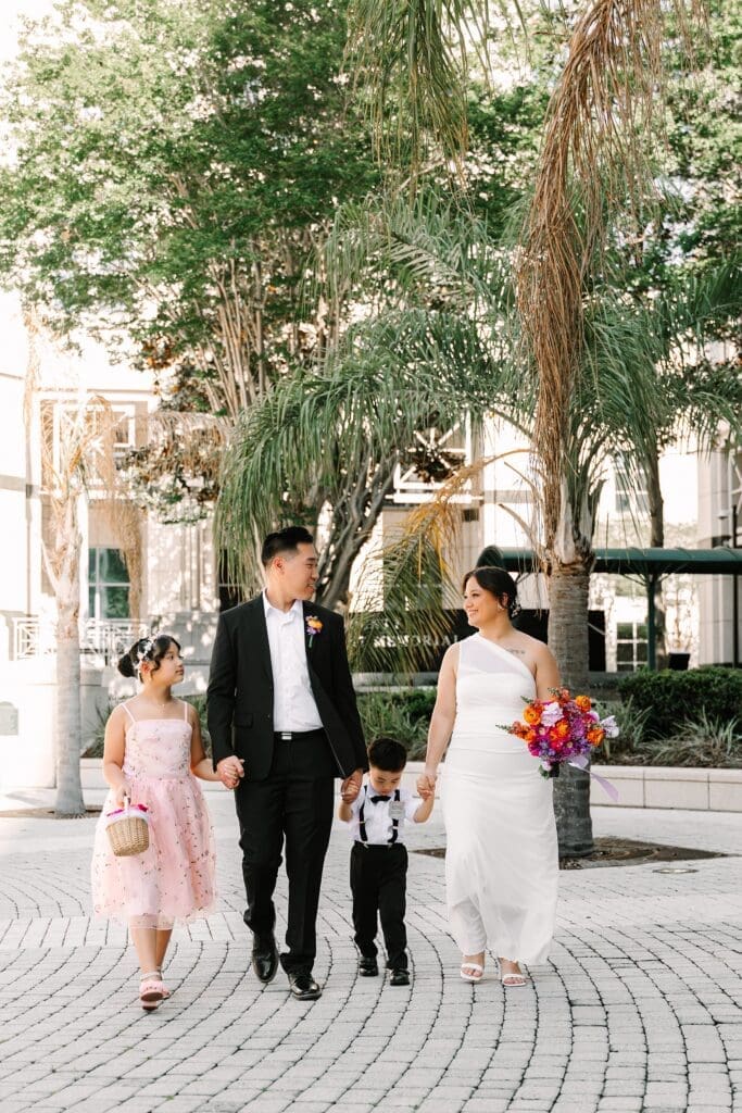 bride, groom, and their two children in formal wedding attire and bright spring florals walk holding hands in front of the Orange County Courthouse in Orlando after their courthouse wedding, captured by Gracie May Photography.
