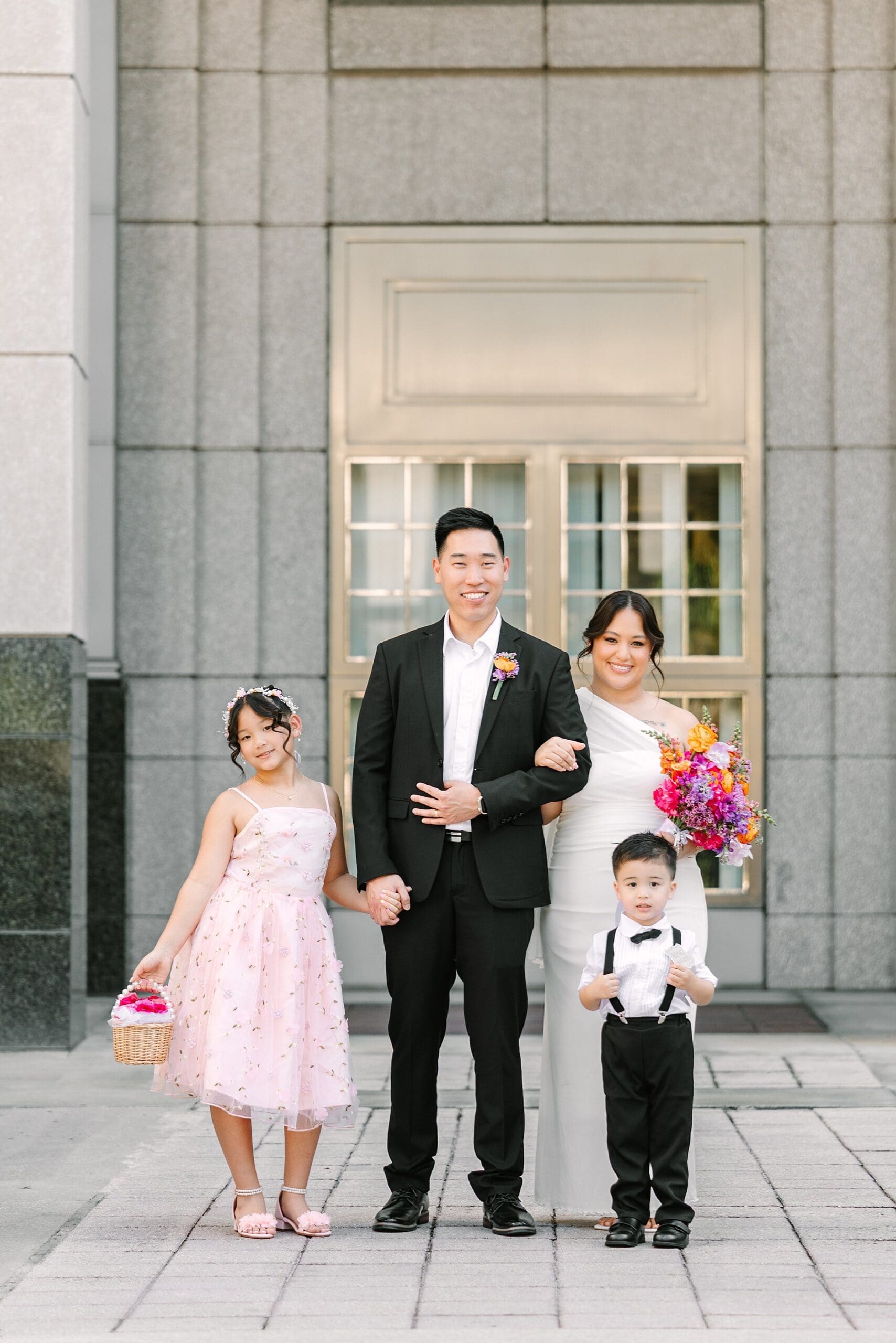 bride, groom, and their two children in formal wedding attire and bright spring florals smile in front of the Orange County Courthouse in Orlando after their courthouse wedding, captured by Gracie May Photography.