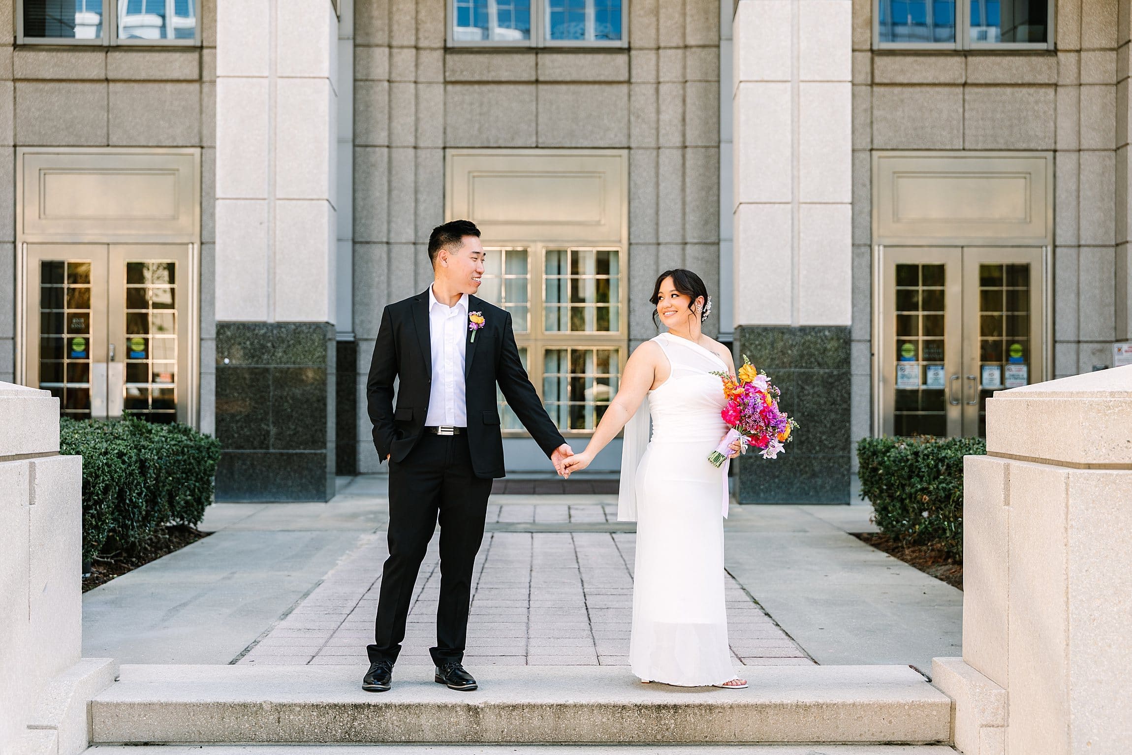 bride and groom in formal wedding attire carrying a bright spring bouquet hold hands outside the Orange County Courthouse in Orlando after their courthouse wedding, captured by Gracie May Photography.