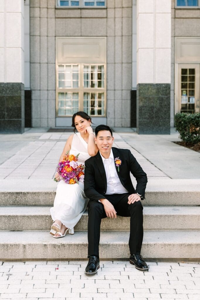 bride and groom in formal wedding attire carrying a bright spring bouquet sit on the steps of the Orange County Courthouse in Orlando after their courthouse wedding, captured by Gracie May Photography.