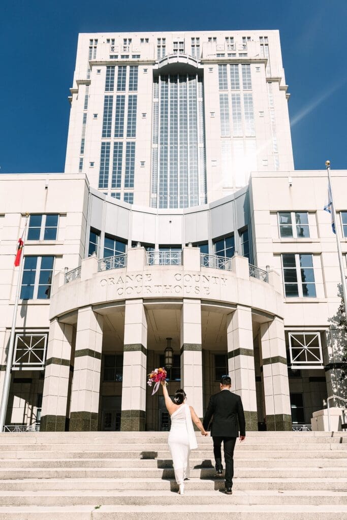 bride and groom in formal wedding attire carrying a bright spring bouquet walk towards the Orange County Courthouse in Orlando after their courthouse wedding, captured by Gracie May Photography.
