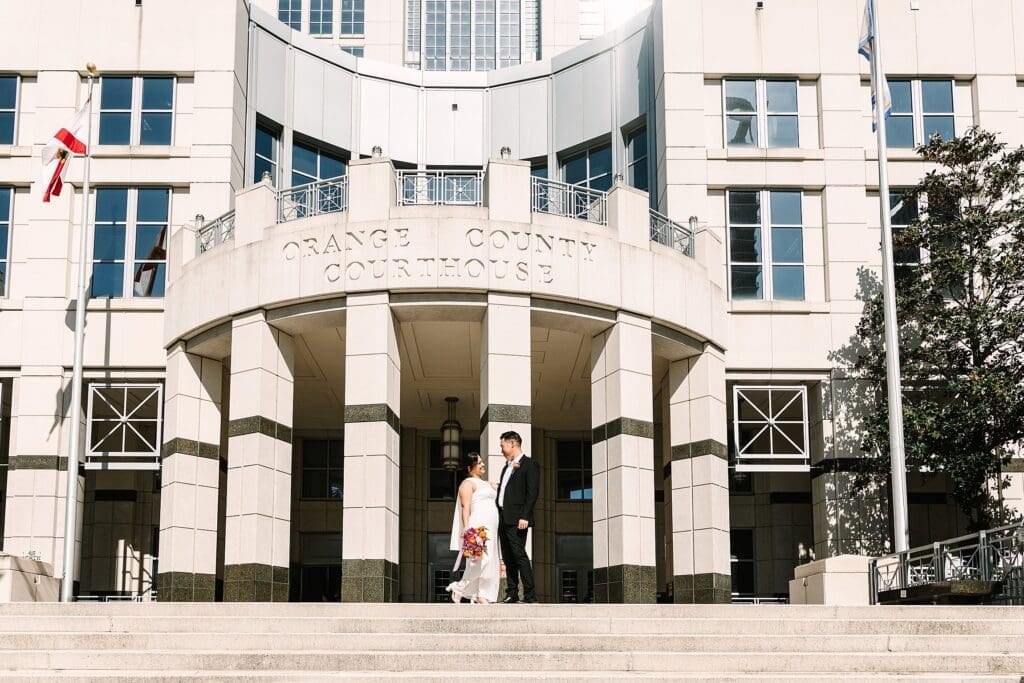 bride and groom in formal wedding attire carrying a bright spring bouquet in front of the Orange County Courthouse in Orlando after their courthouse wedding, captured by Gracie May Photography.