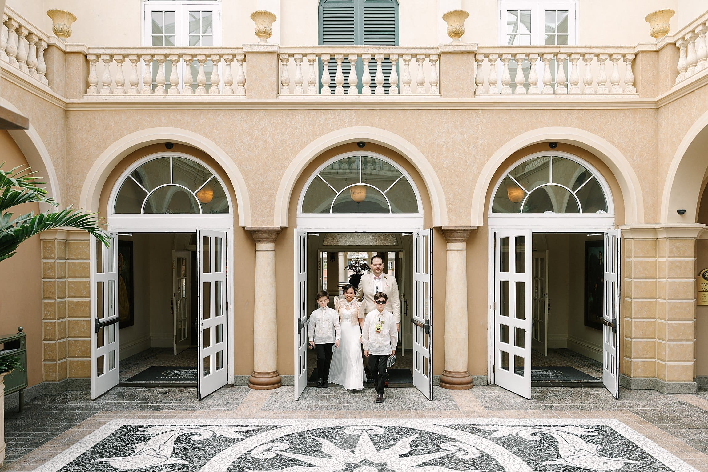 bride in a white dress with a Filipino style jacket and groom in a tan linen suit walk out of the Loews Portofino Bay Hotel at Universal Orlando with their kids, captured by Gracie May Photography