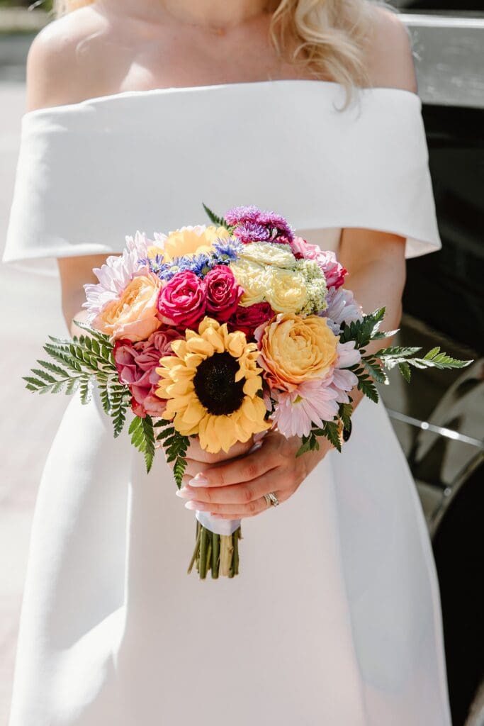 closeup of a bride holding a bridal bouquet with sunflowers and other bright flowers in front of the Osceola Country Courthouse for her courthouse wedding, captured by Gracie May Photography.