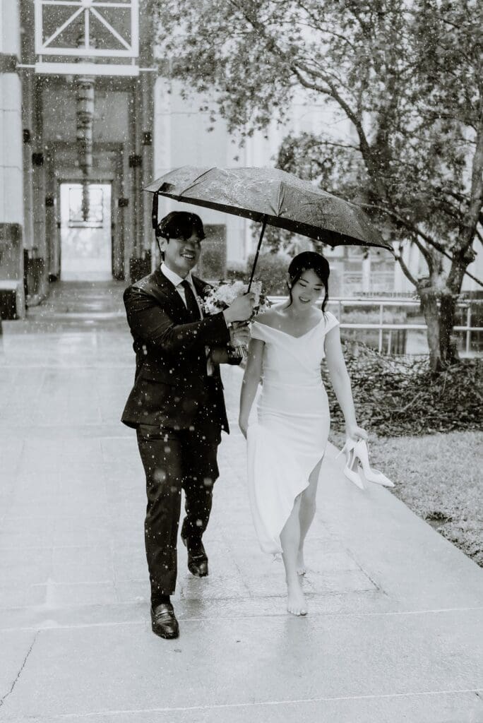 black and white editorial shot of a bride and groom walking in the rain with an umbrella outside the Orlando Orange County Courthouse, captured by Gracie May Photography