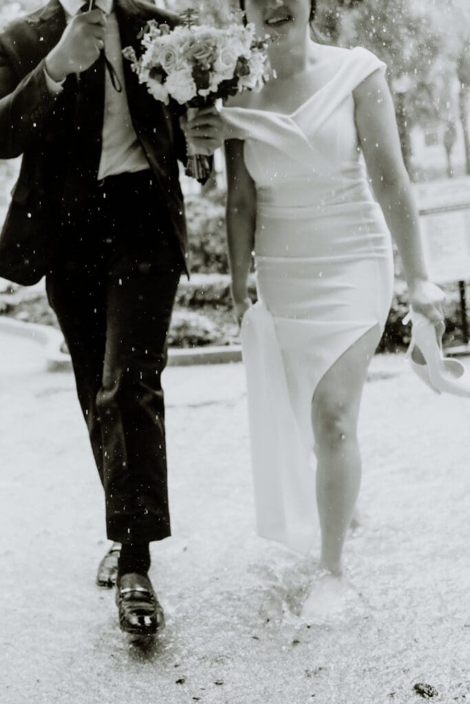 black and white editorial shot of a bride and groom walking in the rain with an umbrella outside the Orlando Orange County Courthouse, captured by Gracie May Photography