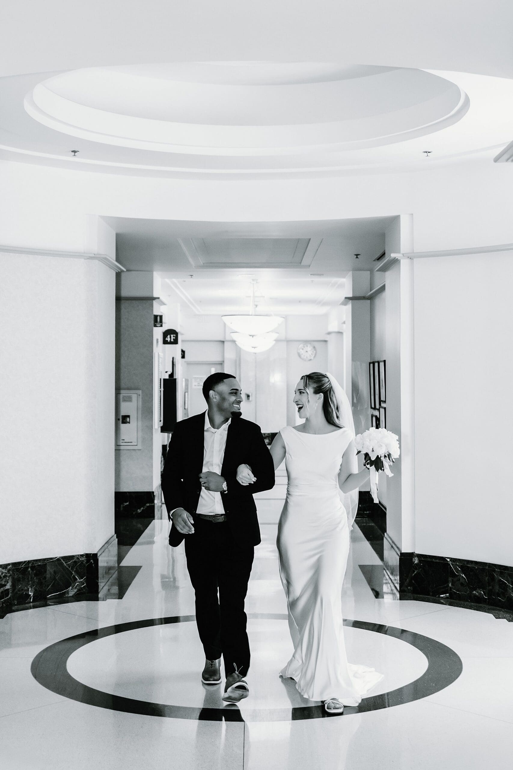 Bride and groom walking arm in arm and laughing together down a curved fourth-floor hallway with a circular medallion in the floor at the Orange County Courthouse in Orlando, photographed in high-contrast black and white.