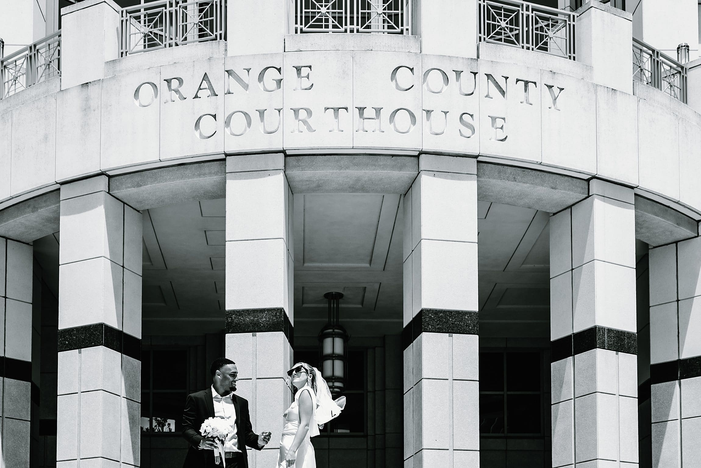 Bride in sunglasses with her veil catching the wind laughing beside the groom, who holds her white bouquet, beneath the engraved Orange County Courthouse lettering at their editorial black and white Orlando courthouse wedding